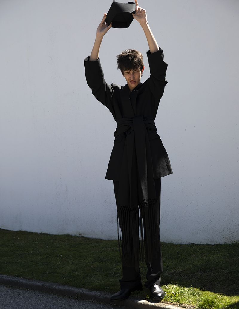 Model wearing a black belted coat lifting a cap against a white wall and curb for L'Officiel Hommes Malaysia