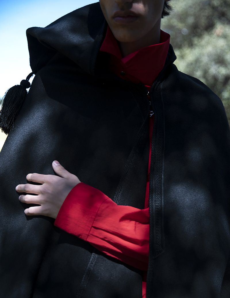 Model wearing a black hooded cape over a vibrant red shirt outdoors in dappled sunlight.