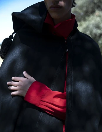 Model wearing a black hooded cape over a vibrant red shirt outdoors in dappled sunlight.