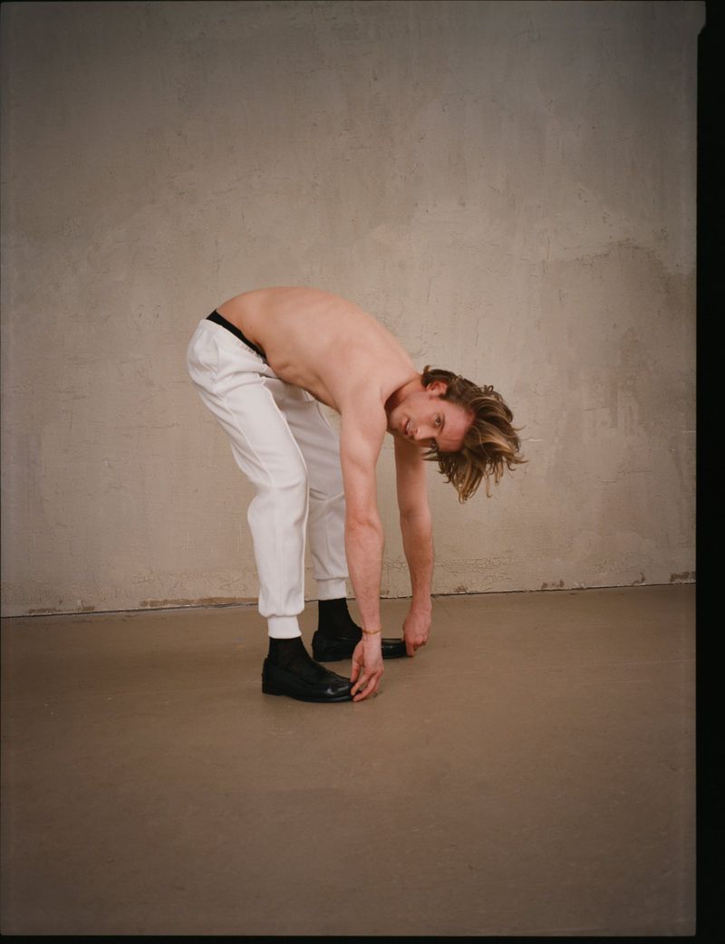 Shirtless male model wearing white trousers and black shoes bending forward in front of a textured wall