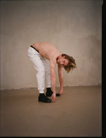 Shirtless male model wearing white trousers and black shoes bending forward in front of a textured wall