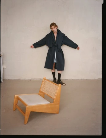 Model RJ King in a dark oversized coat stands against a plain wall behind a wooden lounge chair for The Greatest Magazine.