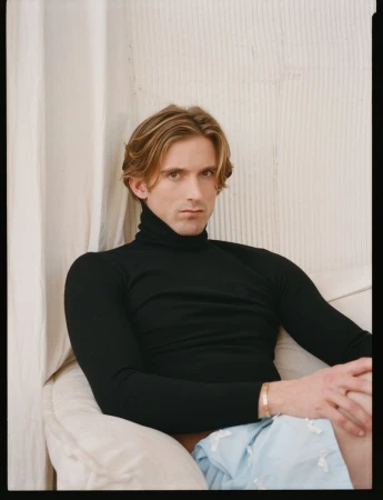 Man wearing a black turtleneck sweater and light blue shorts seated on an off-white chair indoors