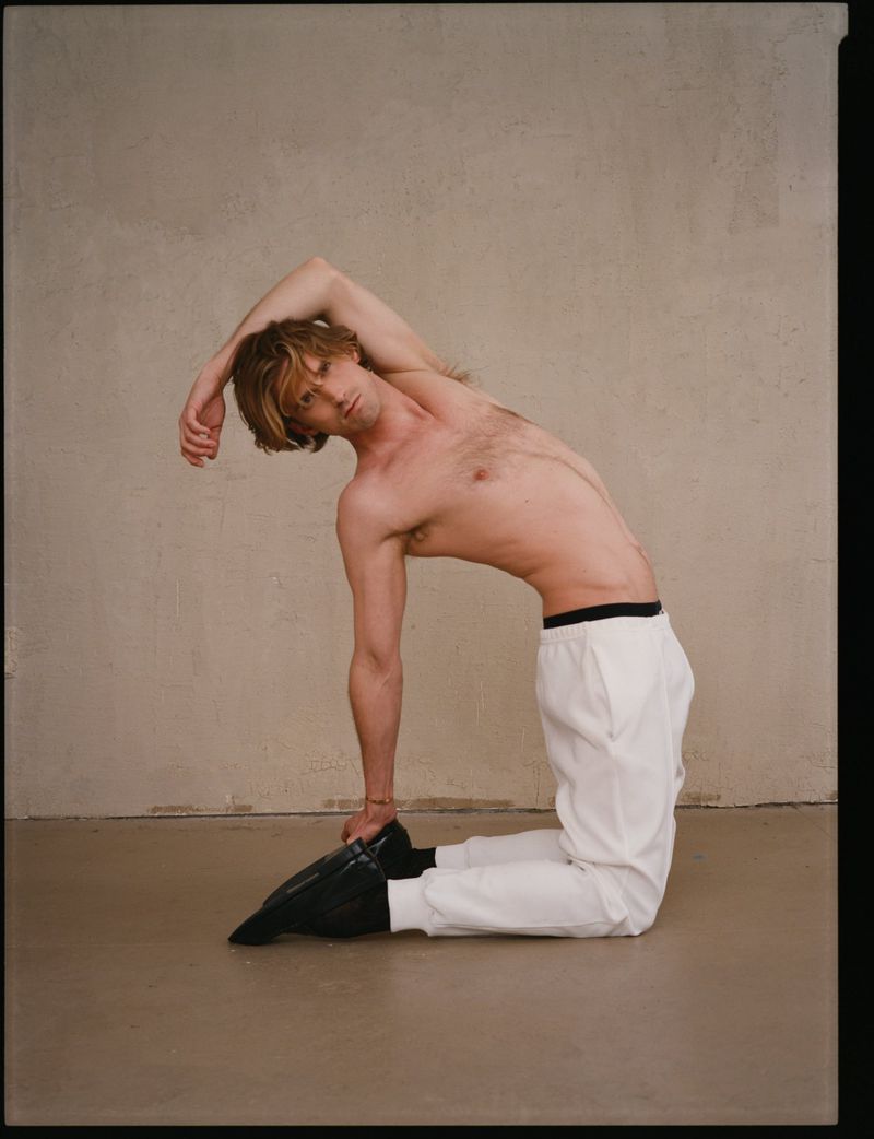 Shirtless male model kneeling and bending backward against a plain neutral wall for The Greatest Magazine editorial