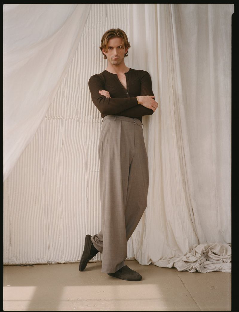 Model RJ King in a dark long-sleeve henley shirt and grey trousers leaning against white draped fabric.
