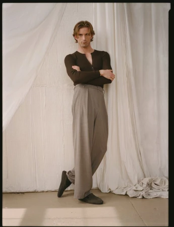 Model RJ King in a dark long-sleeve henley shirt and grey trousers leaning against white draped fabric.