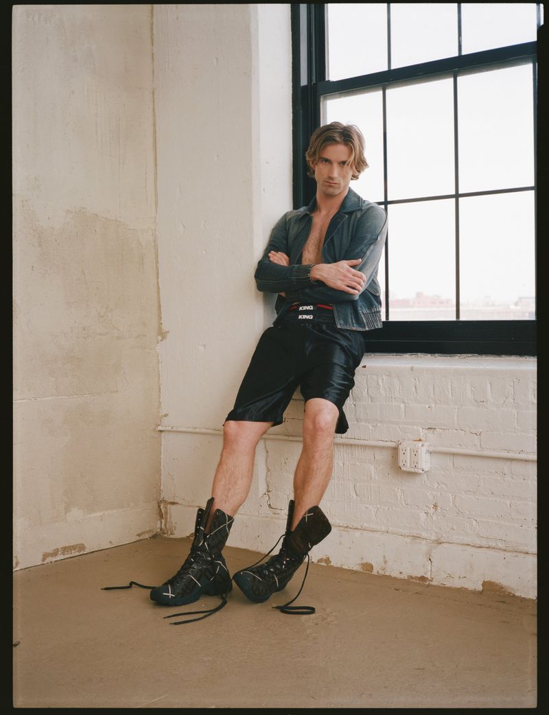 Model RJ King leaning against a multi-pane window wearing a denim jacket and black shorts for The Greatest Magazine.