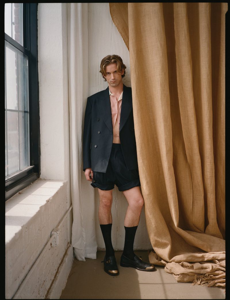 Model RJ King in a dark double-breasted blazer and shorts standing beside a tall window and heavy beige curtain.