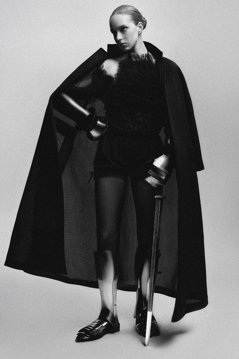 Model in black coat wearing metallic armor greaves and gauntlets holding a baton against a seamless grey backdrop
