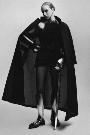 Model in black coat wearing metallic armor greaves and gauntlets holding a baton against a seamless grey backdrop