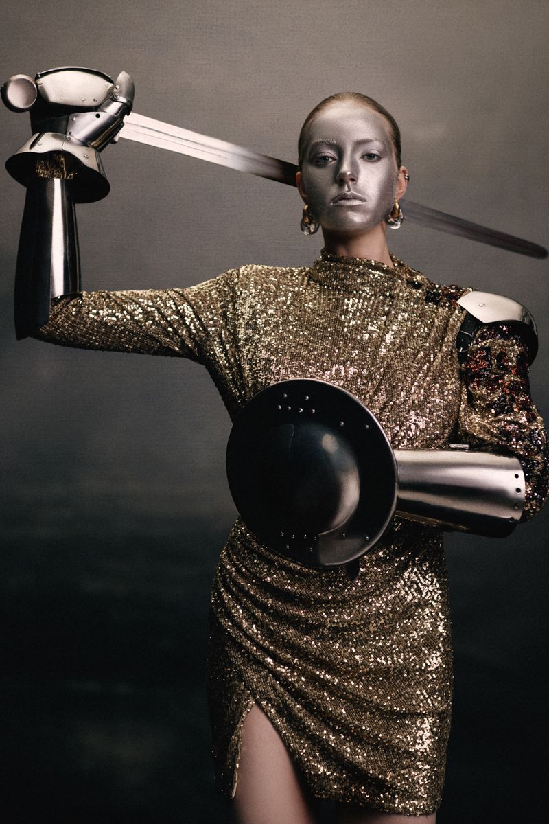 Model with silver painted face wearing gold sequin dress and metallic armor holding a sword for Numéro Switzerland