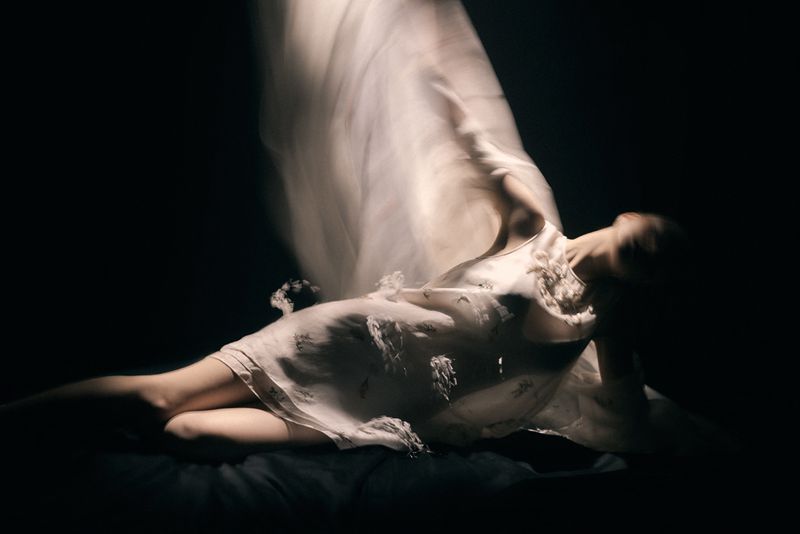 Model in a white dress reclining against a dark background with long exposure motion blur on sheer fabric.