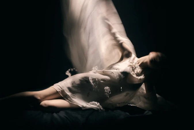 Model in a white dress reclining against a dark background with long exposure motion blur on sheer fabric.