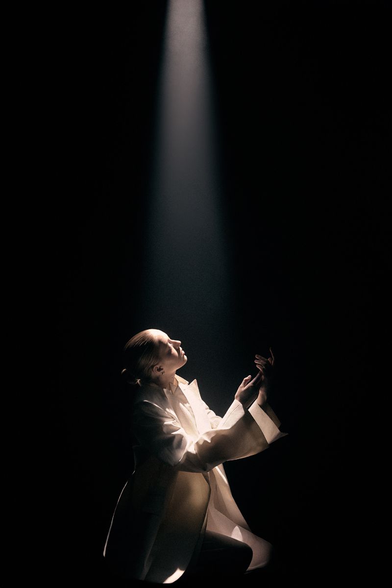 Model kneeling in profile under a dramatic vertical spotlight against a dark background, wearing an ivory white jacket.