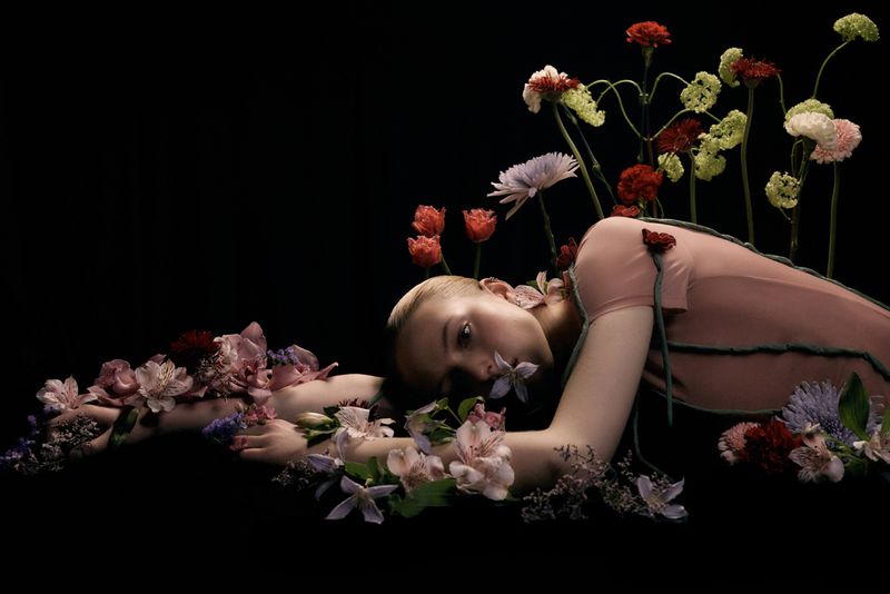 Model lying down surrounded by fresh flowers against a black background for Numéro Switzerland editorial