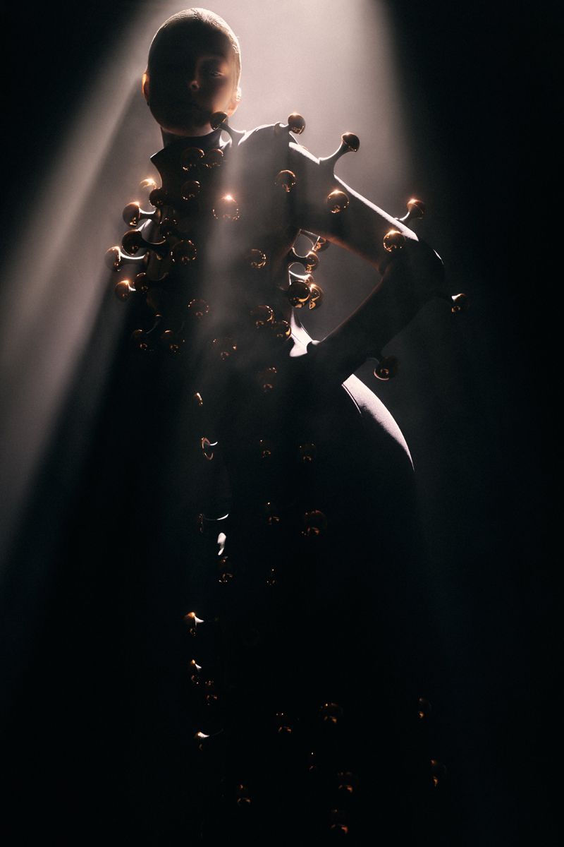 Model in a dark sculptural garment adorned with metallic spherical studs standing in a dramatic beam of light