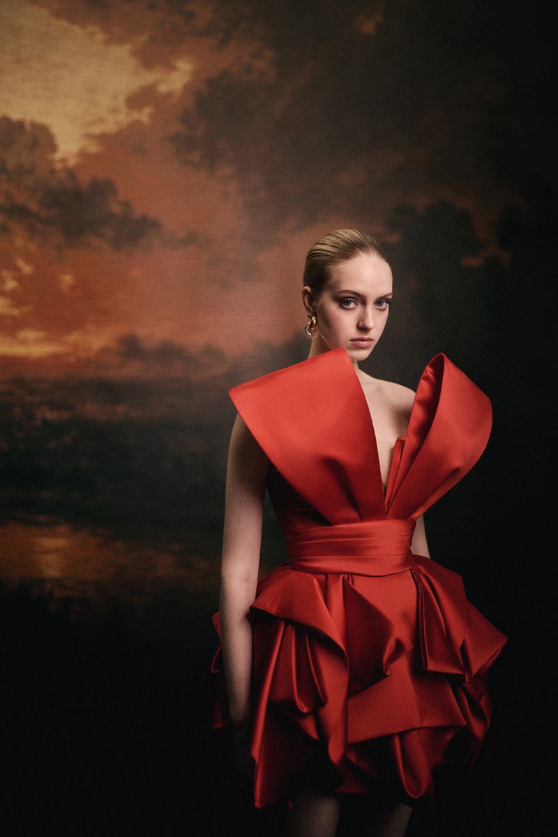 Model in a sculptural red dress with dramatic shoulders against a dark painted cloud backdrop for Numéro Switzerland