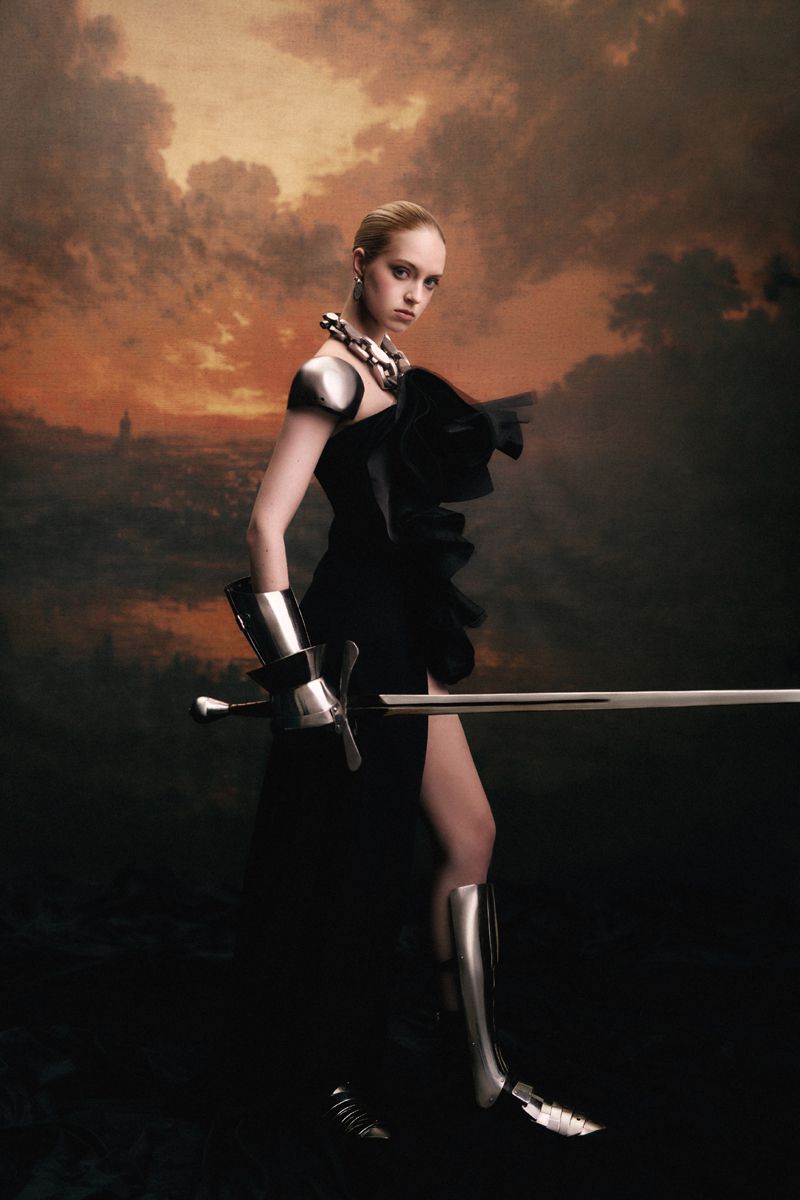 Model wearing a black dress and metallic armor pieces holding a sword against a painted sunset landscape backdrop.