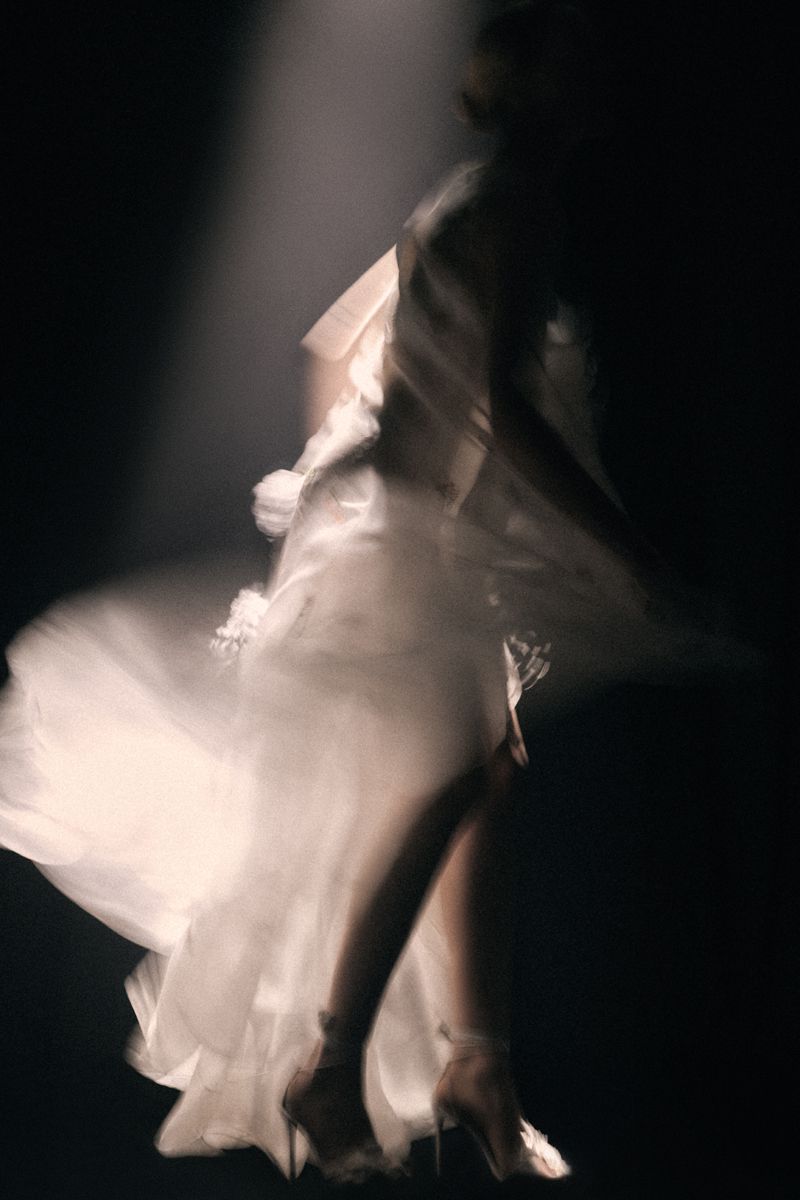 Motion blurred figure of a woman in a flowing white gown captured under a dramatic spotlight against a dark background