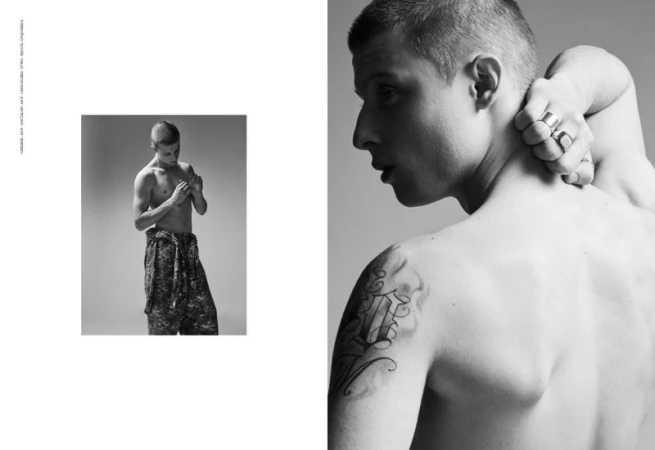 Black and white editorial portrait of a shirtless man with tattoos wearing patterned trousers against a grey backdrop