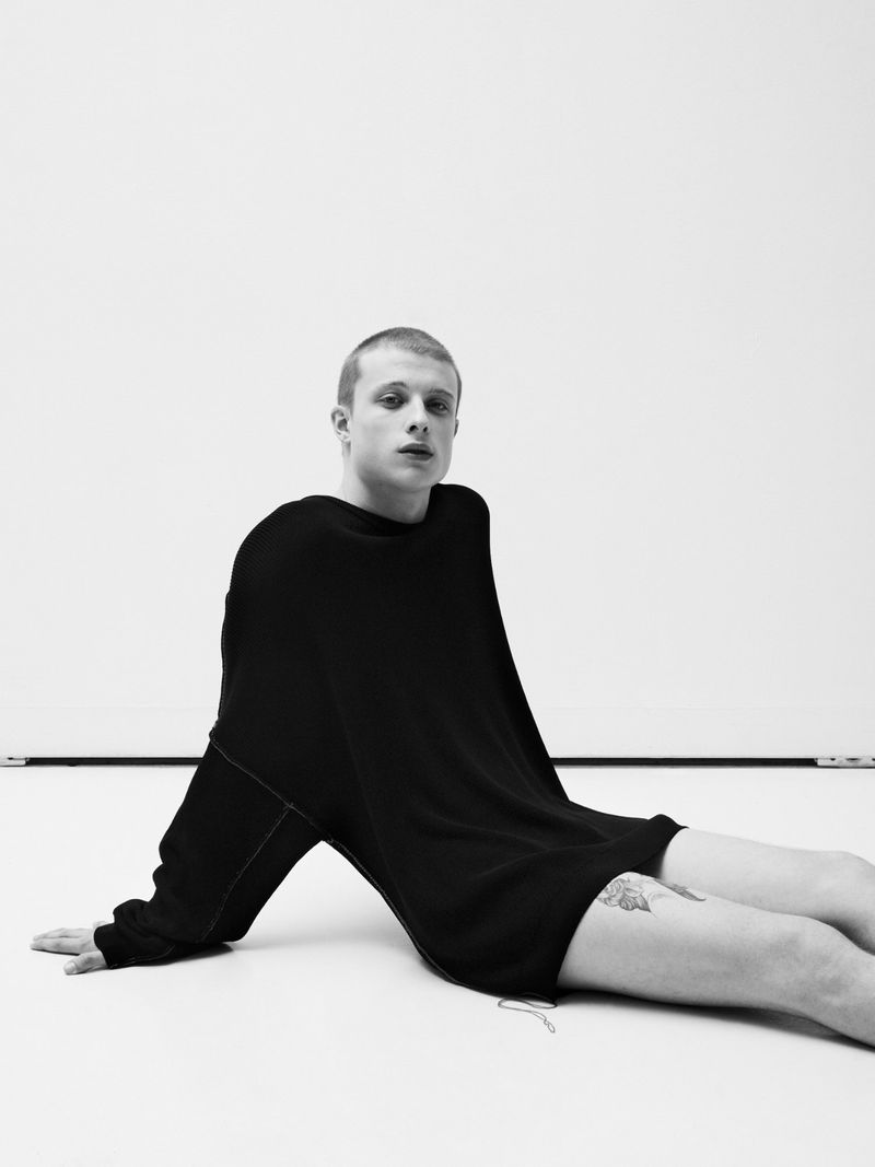 Young man with a buzz cut wearing an oversized black sweater seated against a plain white studio background
