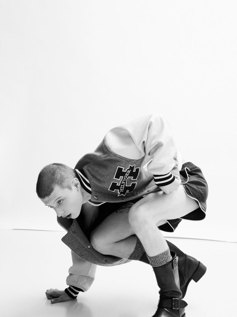 A man in a varsity jacket and shorts crouches on a white studio floor in a black and white editorial image.