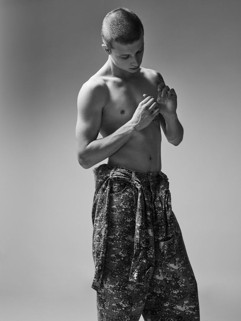 Monochrome portrait of a shirtless young man with short hair wearing patterned trousers against a gradient grey studio background