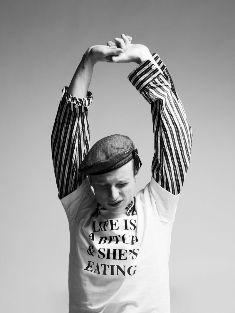 Monochrome portrait of a man in a flat cap and graphic t-shirt stretching his arms upward against a grey background.