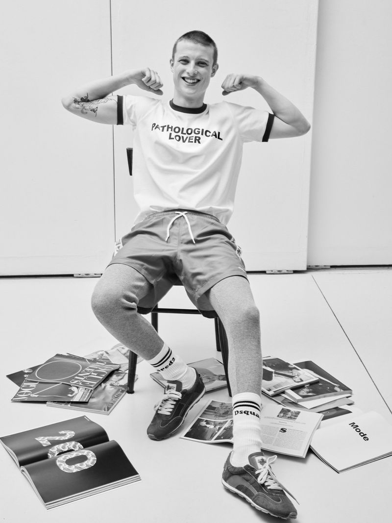Young man with short hair wearing graphic t-shirt and shorts seated on a chair amid scattered magazines in monochrome editorial photograph.