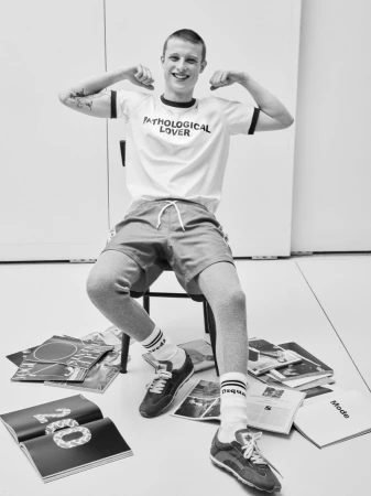 Young man with short hair wearing graphic t-shirt and shorts seated on a chair amid scattered magazines in monochrome editorial photograph.