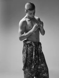 Monochrome portrait of a shirtless young man with short hair wearing patterned trousers against a gradient grey studio background