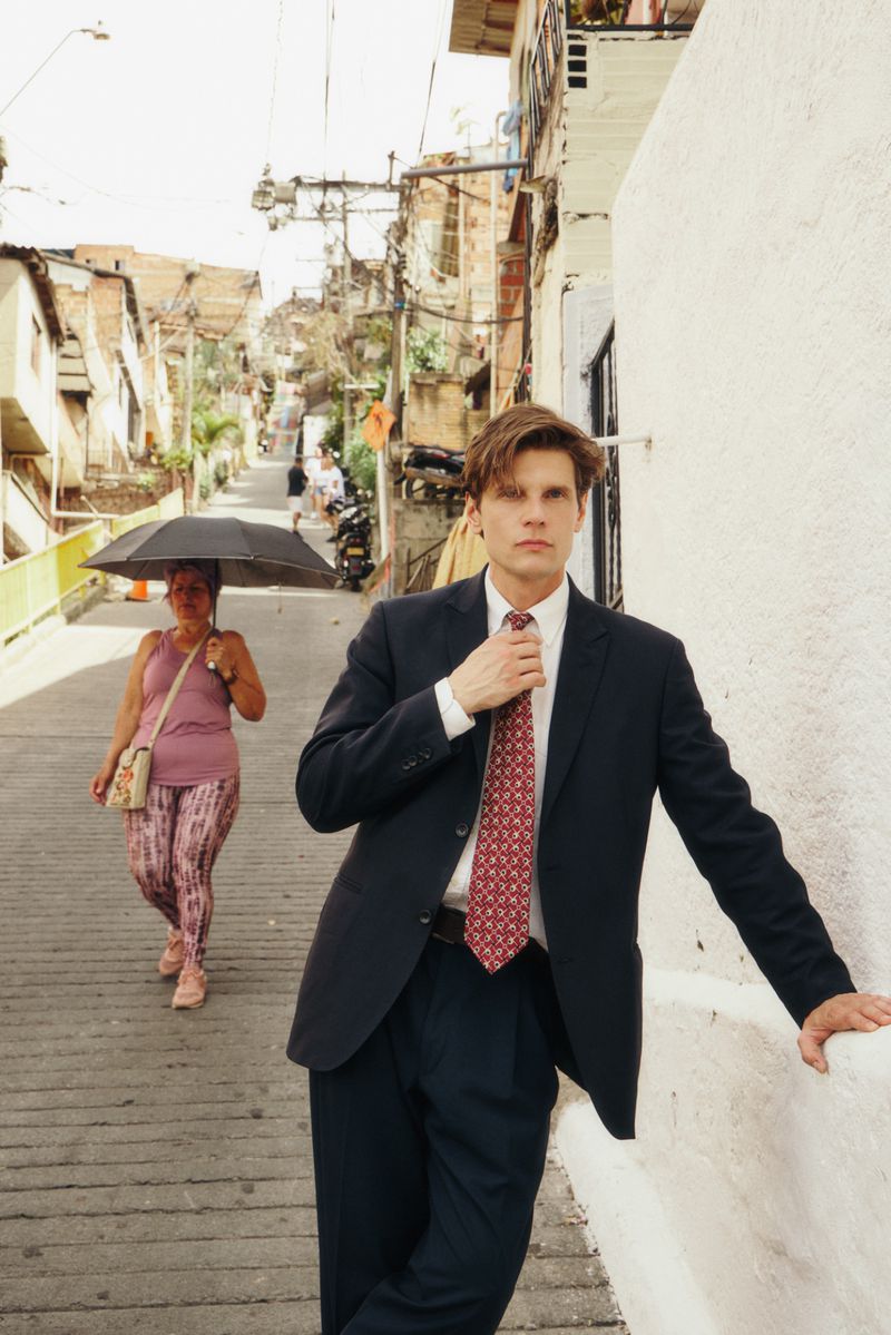 Model in dark suit and patterned red tie leaning against a white wall on a sloping street with a woman walking behind