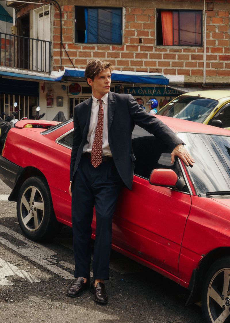 Man wearing a dark suit leaning against a red car on a street in front of brick buildings