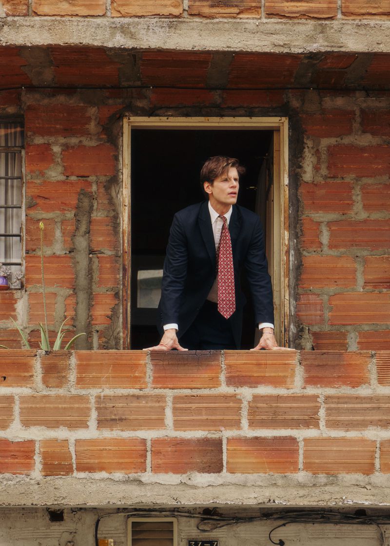 Man wearing a dark suit and patterned red tie leaning out of a doorway on a exposed brick balcony.