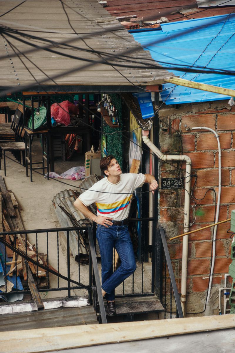 Model Alessandro Egger wearing a striped white t-shirt and blue jeans standing on an outdoor metal staircase in Medellin.