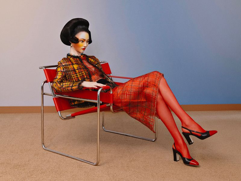 Model seated in a red chrome chair wearing a plaid jacket and skirt with red tights and heels