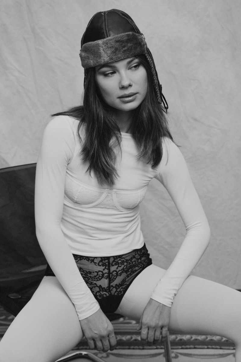 Model seated wearing a leather aviator cap, white long-sleeve top, and lace underwear against a textured backdrop