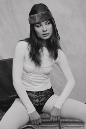 Model seated wearing a leather aviator cap, white long-sleeve top, and lace underwear against a textured backdrop