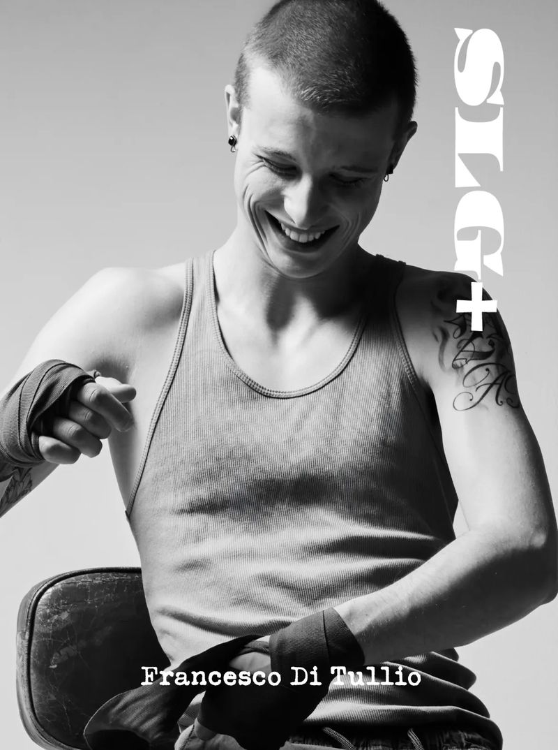 Black and white portrait of a smiling, fair-skinned young man with tattoos and wrapped hands, sitting on a chair, against a plain background.