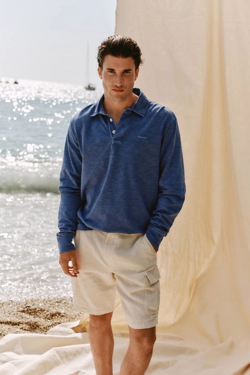 Model wearing a blue polo shirt and beige cargo shorts standing before an ocean view with draped fabric for Gant