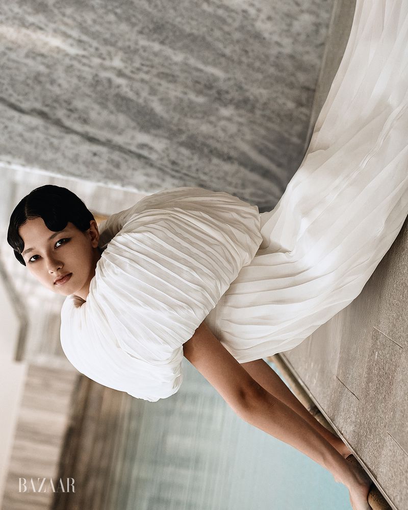 Model wearing a white pleated dress with voluminous sleeves posing against textured stone and water background.