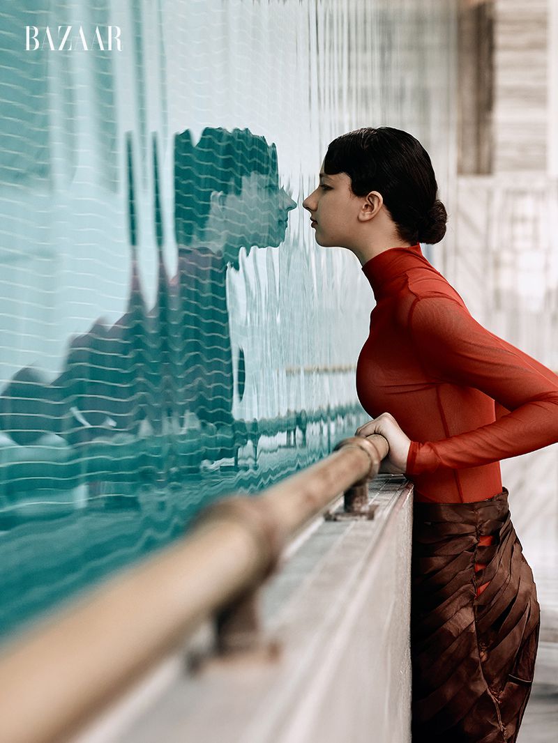 Model in a red sheer turtleneck leaning over a railing looking at her reflection in blue water for Harper's Bazaar