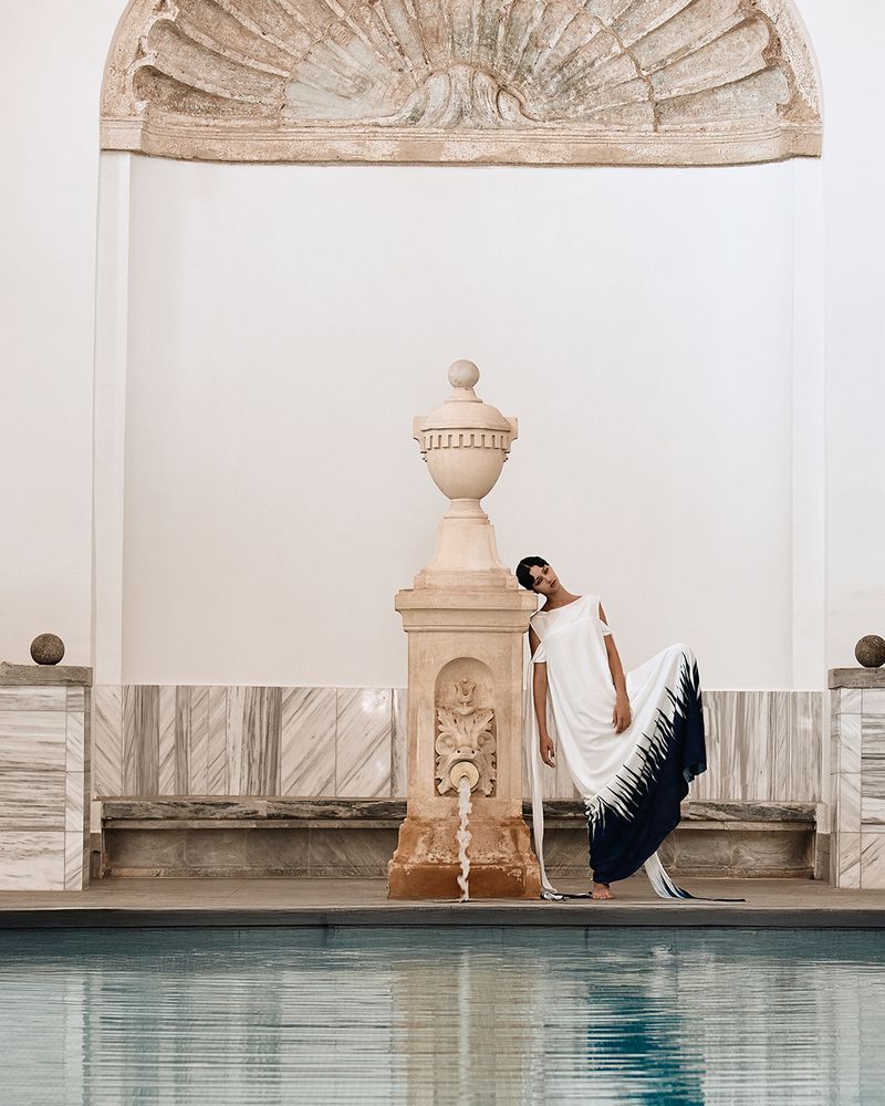 Woman in elegant white dress poses by vintage fountain beside swimming pool in outdoor setting with marble architectural background.