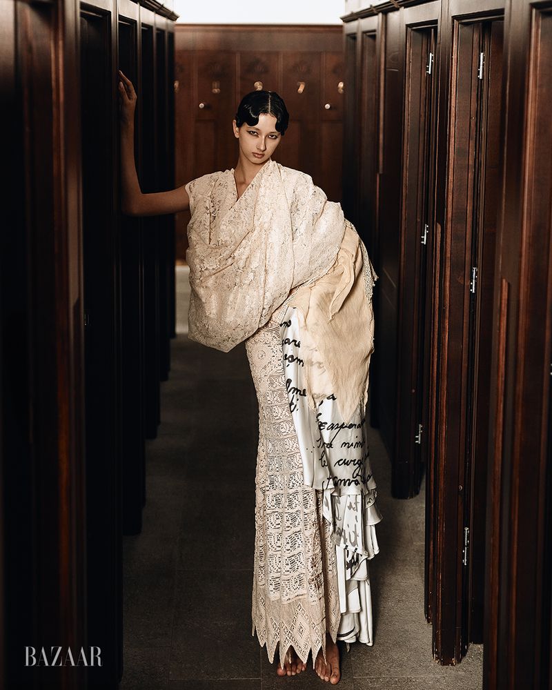 Model wearing a patterned cream lace dress and wrap posing between dark wood lockers in an editorial portrait for Venus.