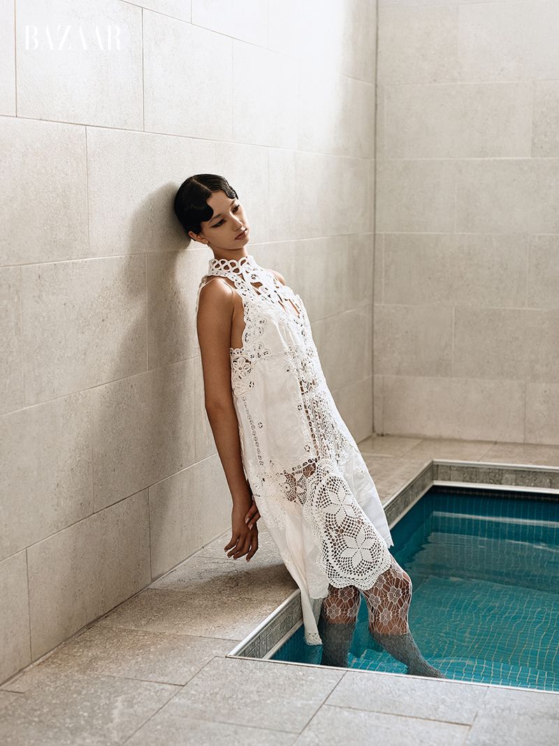 Model in white lace dress leaning against stone wall next to indoor pool for Harper's Bazaar Ecuador editorial
