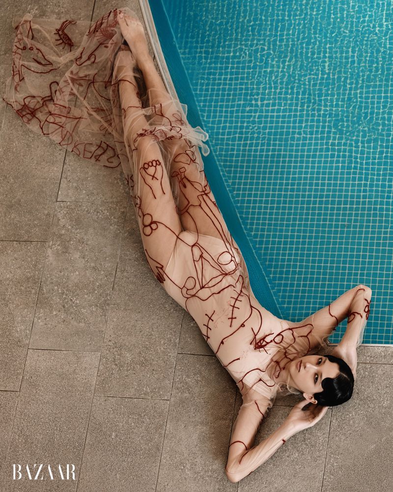 Model lying beside a blue tiled swimming pool wearing a sheer dress with red embroidered line drawings for Harper's Bazaar Ecuador.