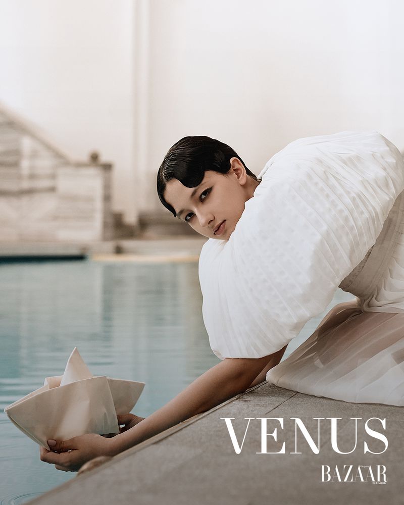 Model in white dress leaning by an outdoor pool holding a paper boat for Harper's Bazaar Ecuador editorial