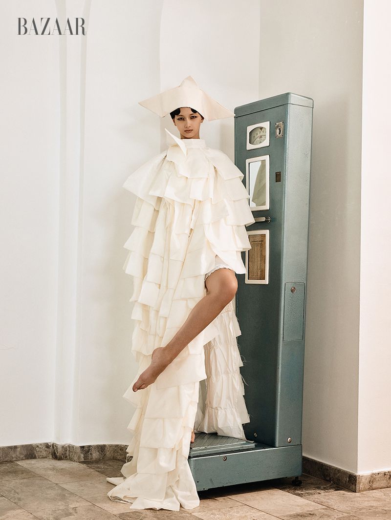 Model wearing a structured white tiered gown and geometric hat standing beside an antique scale in an interior space.