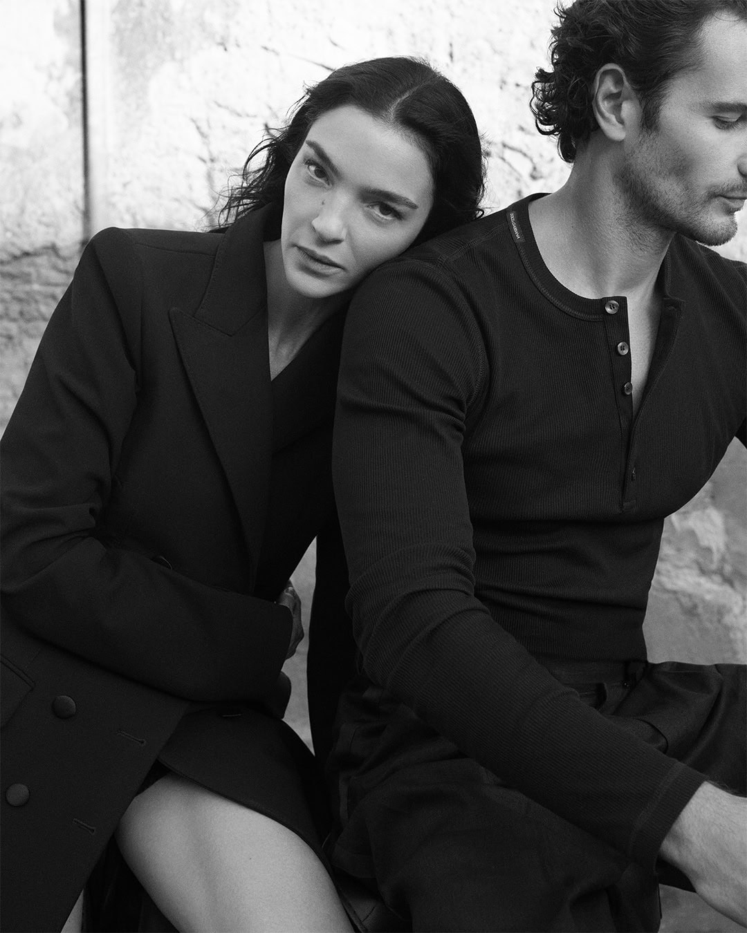Black and white portrait of a woman resting her head on a man's shoulder wearing dark tailored attire for Dolce & Gabbana.