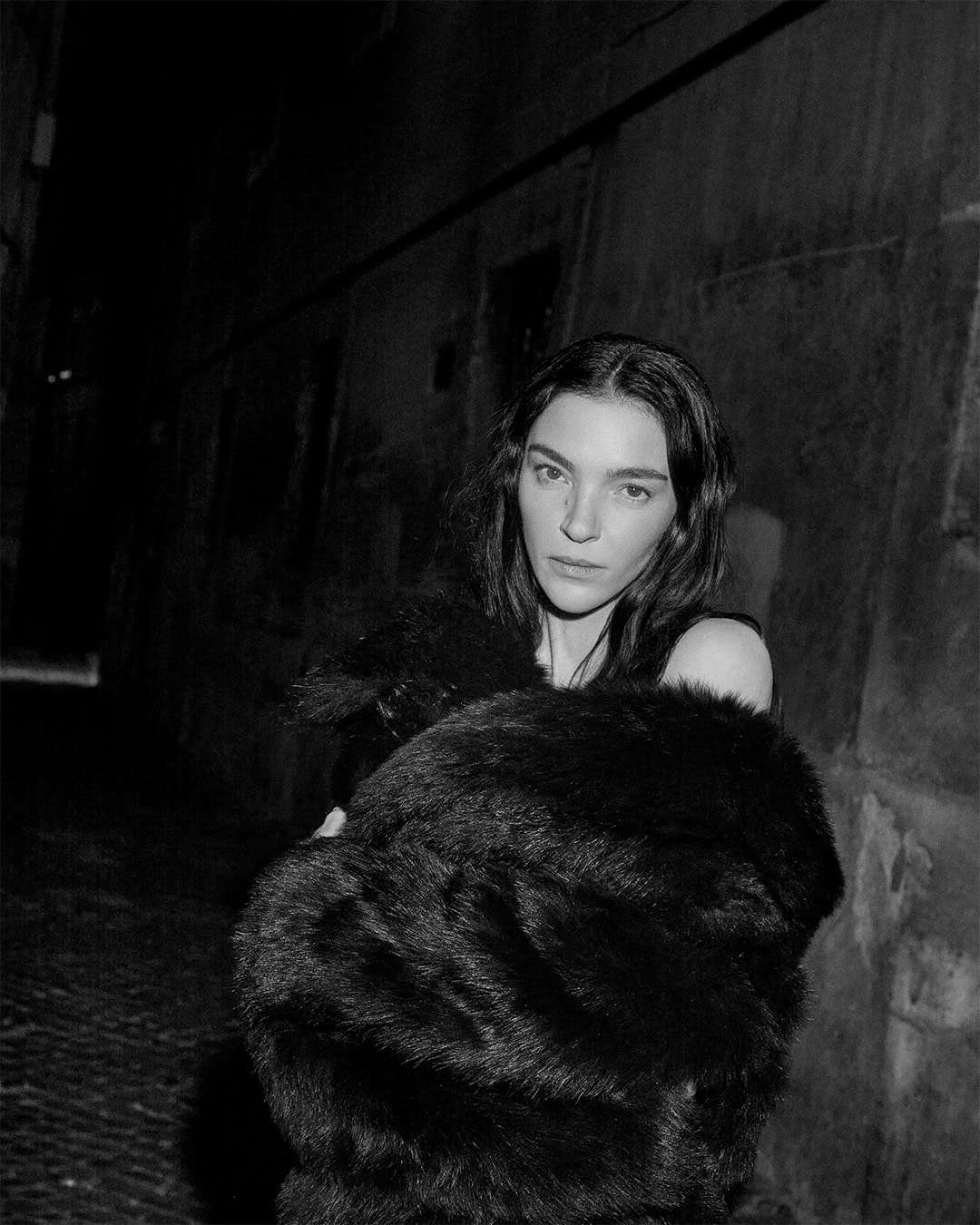 Model Mariacarla Boscono in a dark textured coat standing in a narrow alleyway at night for Dolce & Gabbana